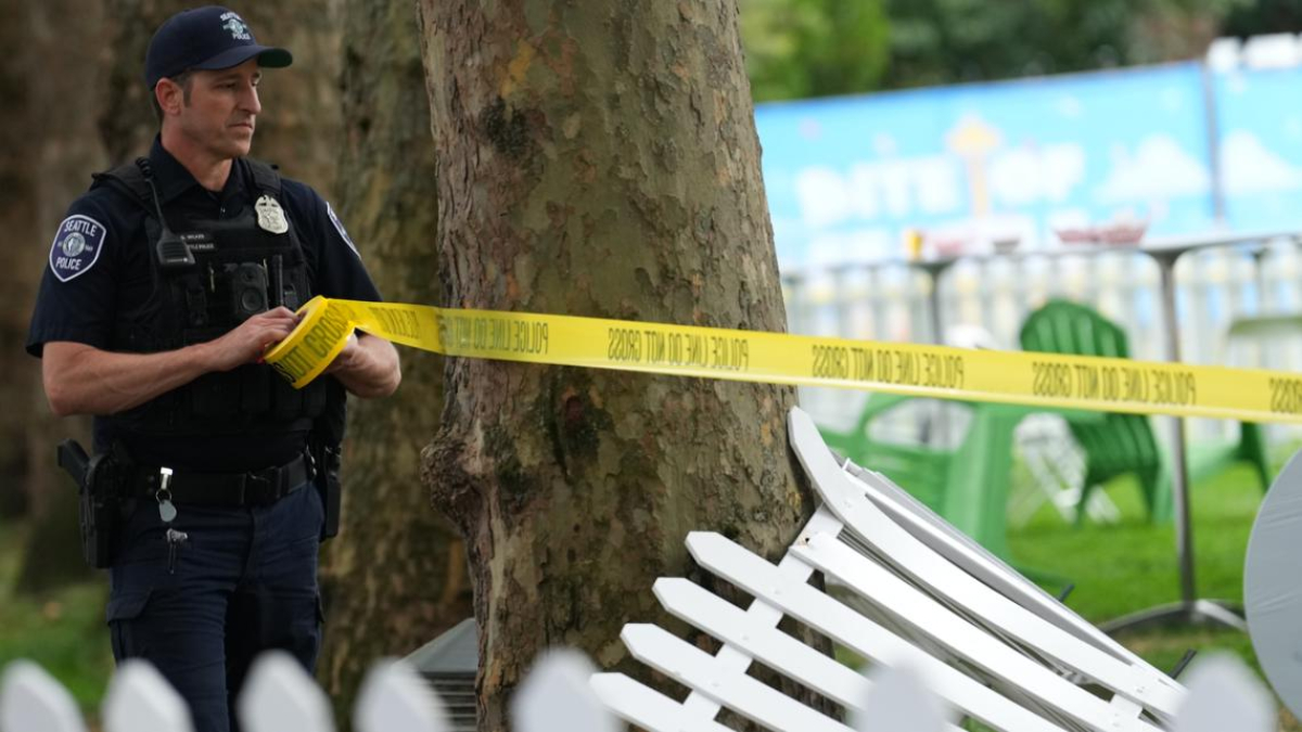 Police officer standing behind yellow caution tape at a park, securing a crime-scene area near a tree and fence pieces ahead of blue banners in the background.