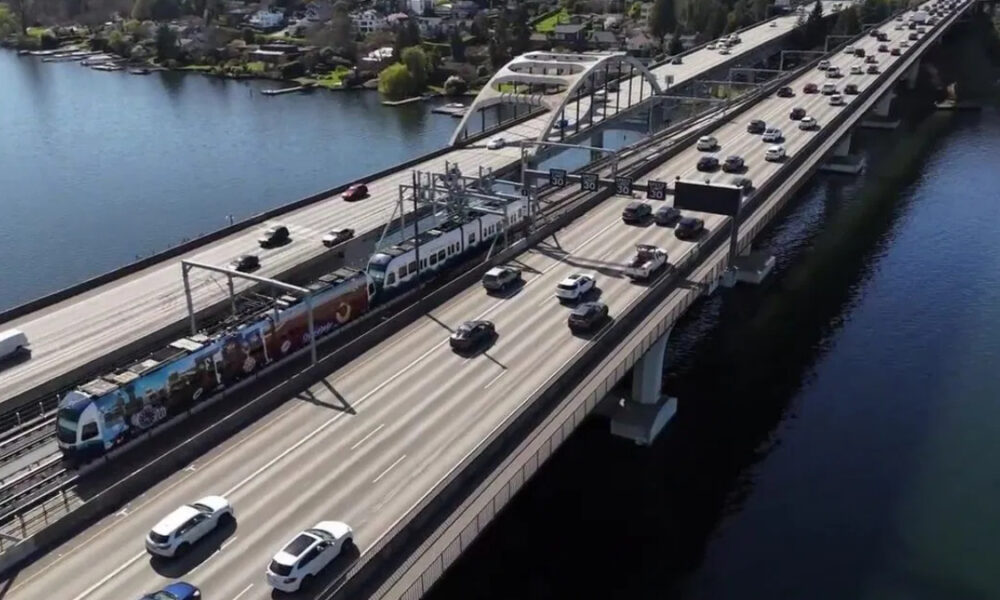 Aerial view of a multi-lane bridge over a river with a passenger train on the tracks and heavy traffic on both sides of the bridge.