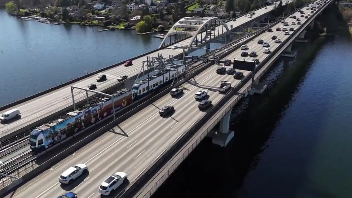Aerial view of a multi-lane bridge over a river with a passenger train on the tracks and heavy traffic on both sides of the bridge.