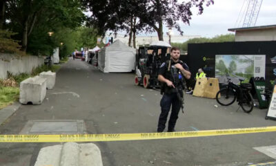 Police officer in tactical gear stands behind yellow caution tape guarding a cordoned-off street with white tents and temporary setup behind him.