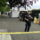 Police officer in tactical gear stands behind yellow caution tape guarding a cordoned-off street with white tents and temporary setup behind him.