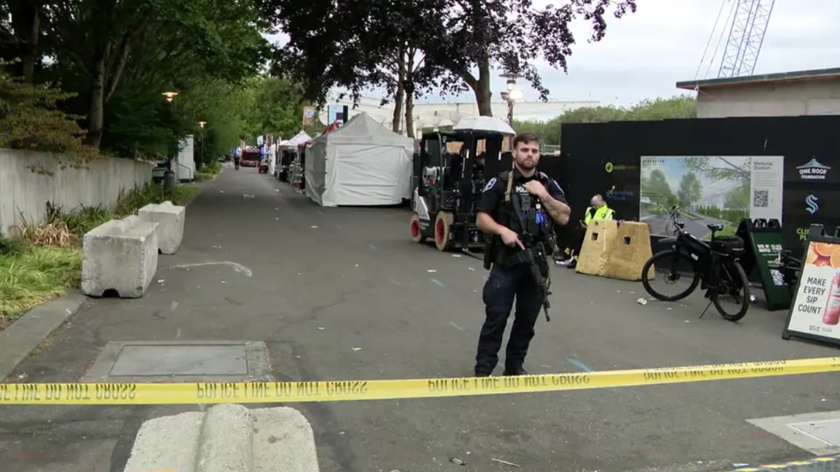 Police officer in tactical gear stands behind yellow caution tape guarding a cordoned-off street with white tents and temporary setup behind him.