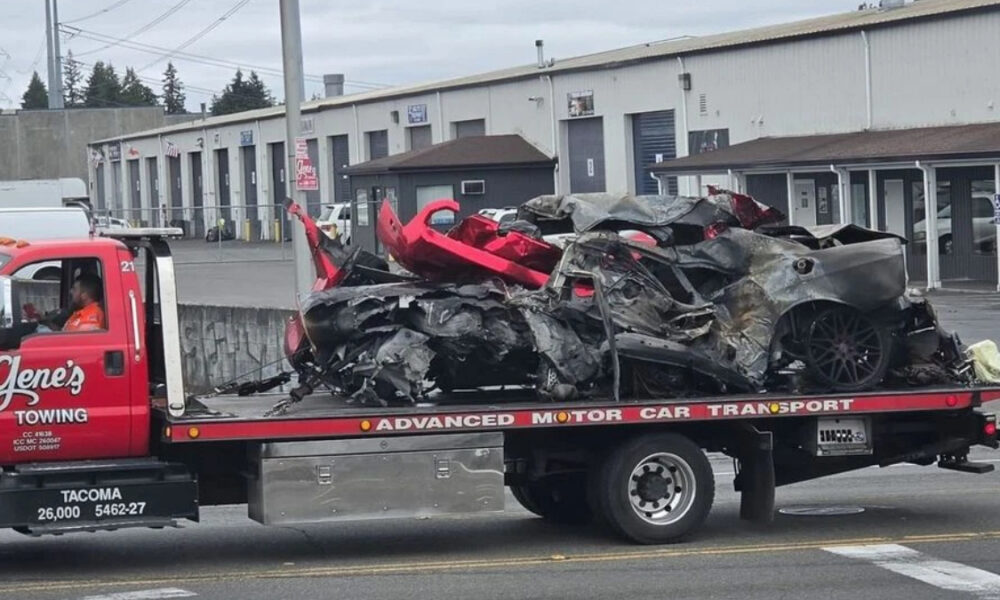 Tow truck carrying a severely wrecked gray sports car on a flatbed in an industrial lot