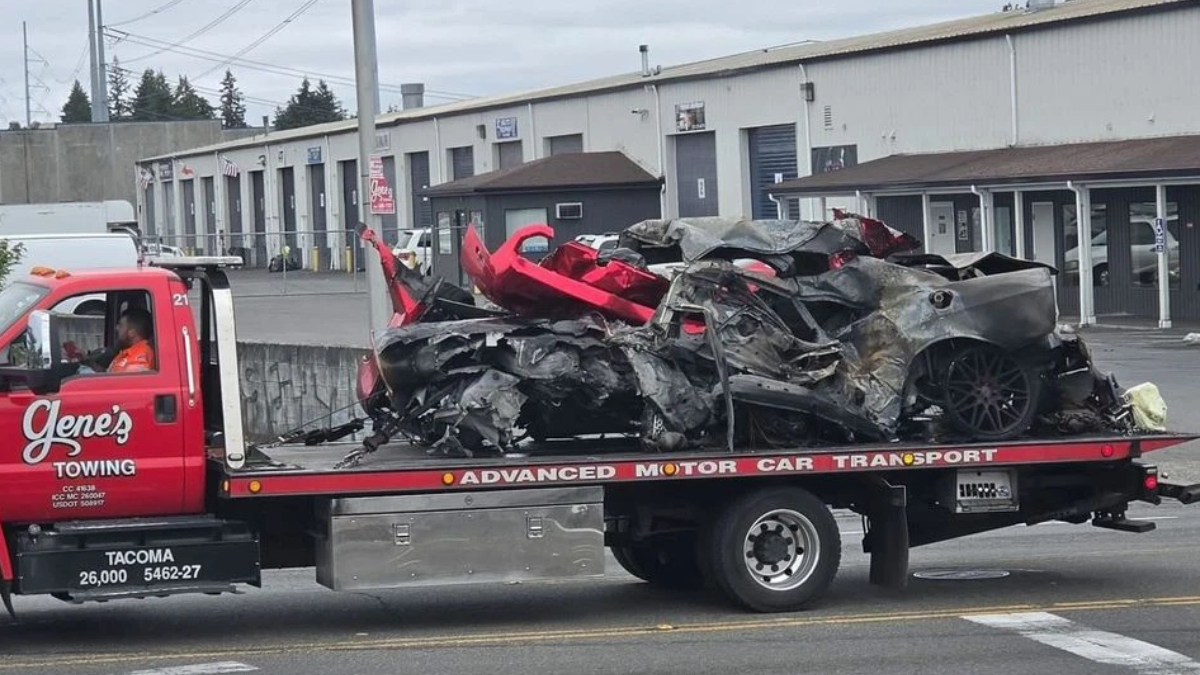 Tow truck carrying a severely wrecked gray sports car on a flatbed in an industrial lot
