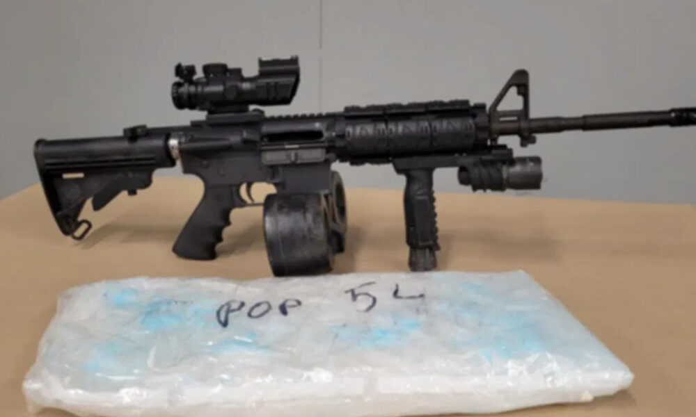 Black rifle with a scope and foregrip on a table, placed beside a blue-wrapped package in the foreground.