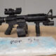 Black rifle with a scope and foregrip on a table, placed beside a blue-wrapped package in the foreground.