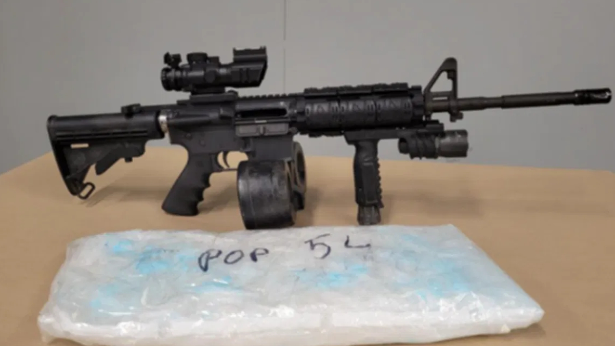 Black rifle with a scope and foregrip on a table, placed beside a blue-wrapped package in the foreground.