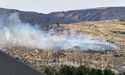 Smoke from a brush fire on a dry hillside behind a suburban neighborhood, with utility poles visible in the foreground.