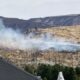 Smoke from a brush fire on a dry hillside behind a suburban neighborhood, with utility poles visible in the foreground.