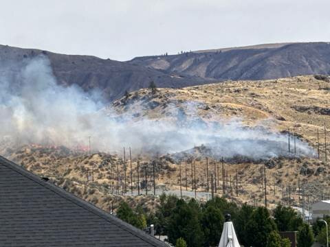Smoke from a brush fire on a dry hillside behind a suburban neighborhood, with utility poles visible in the foreground.