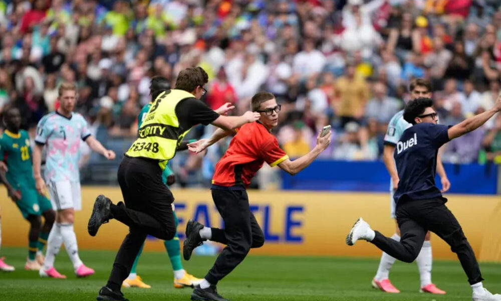 Security guard in a yellow vest chases a man in red who is taking a selfie on a soccer field as players run nearby.