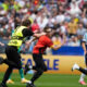 Security guard in a yellow vest chases a man in red who is taking a selfie on a soccer field as players run nearby.