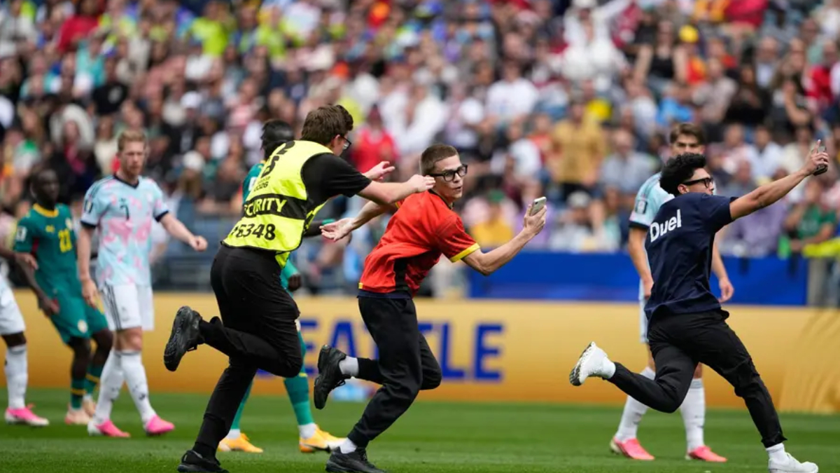 Security guard in a yellow vest chases a man in red who is taking a selfie on a soccer field as players run nearby.