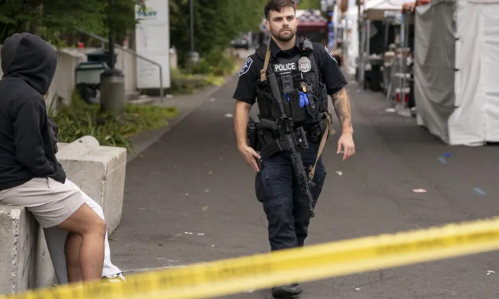 Police officer in tactical gear walking past a hooded person sitting by a concrete barrier, with yellow caution tape in the foreground at an outdoor street area.