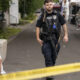 Police officer in tactical gear walking past a hooded person sitting by a concrete barrier, with yellow caution tape in the foreground at an outdoor street area.