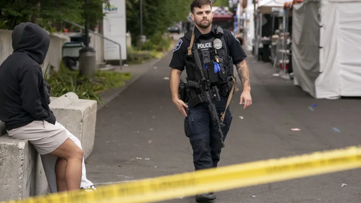 Police officer in tactical gear walking past a hooded person sitting by a concrete barrier, with yellow caution tape in the foreground at an outdoor street area.