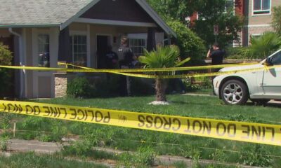 Residential front-yard crime scene with officers behind yellow 'Police Line Do Not Cross' tape blocking entry to the house.