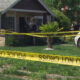 Residential front-yard crime scene with officers behind yellow 'Police Line Do Not Cross' tape blocking entry to the house.