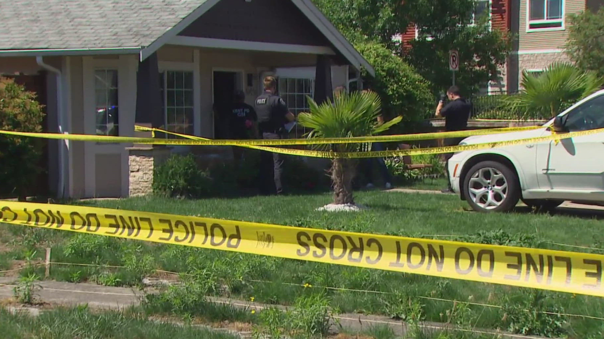 Residential front-yard crime scene with officers behind yellow 'Police Line Do Not Cross' tape blocking entry to the house.