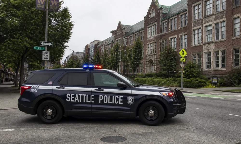 Seattle Police SUV with red and blue lights parked on a tree-lined city street in front of brick, campus-style buildings.