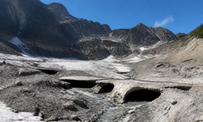 Snowy rocky basin with dark icicle holes and meltwater stream under a blue sky, surrounded by jagged mountains.