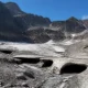 Snowy rocky basin with dark icicle holes and meltwater stream under a blue sky, surrounded by jagged mountains.
