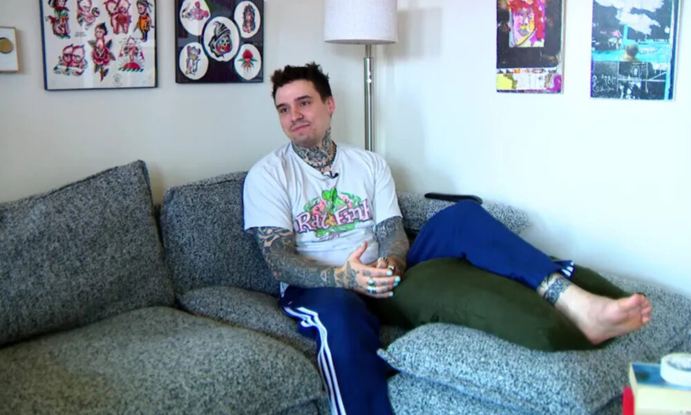 Tattooed man in a Rat Fink T-shirt relaxing on a gray sofa in a living room setting.