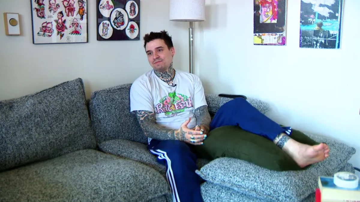 Tattooed man in a Rat Fink T-shirt relaxing on a gray sofa in a living room setting.