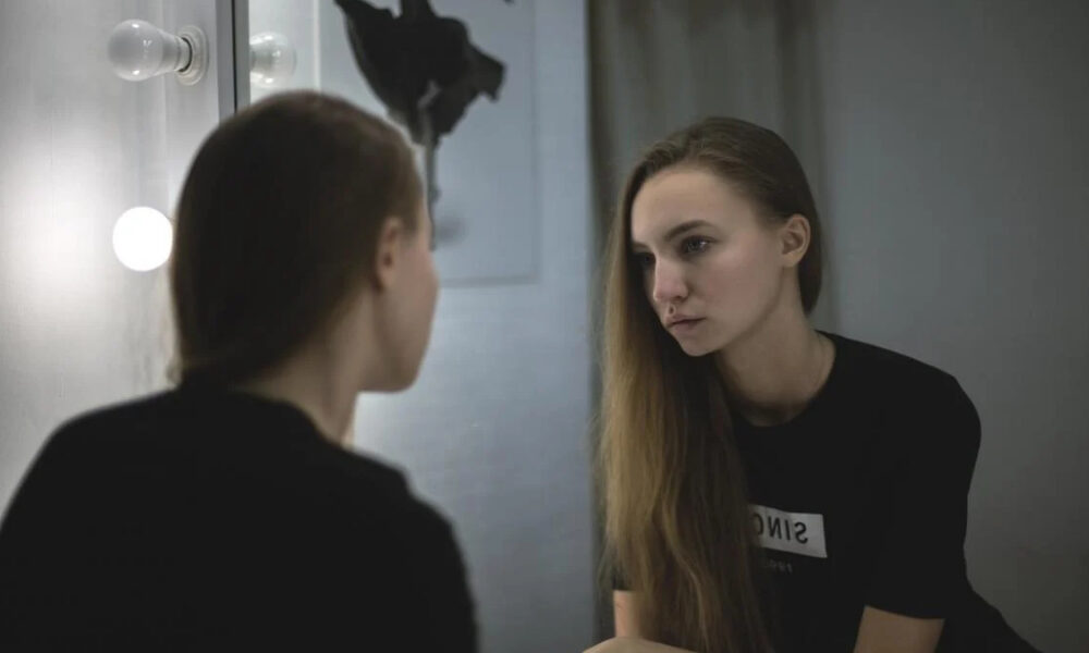 Young woman in a black shirt sits by a vanity mirror with round bulbs, gazing at her reflection.