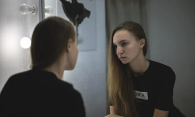 Young woman in a black shirt sits by a vanity mirror with round bulbs, gazing at her reflection.