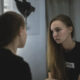 Young woman in a black shirt sits by a vanity mirror with round bulbs, gazing at her reflection.