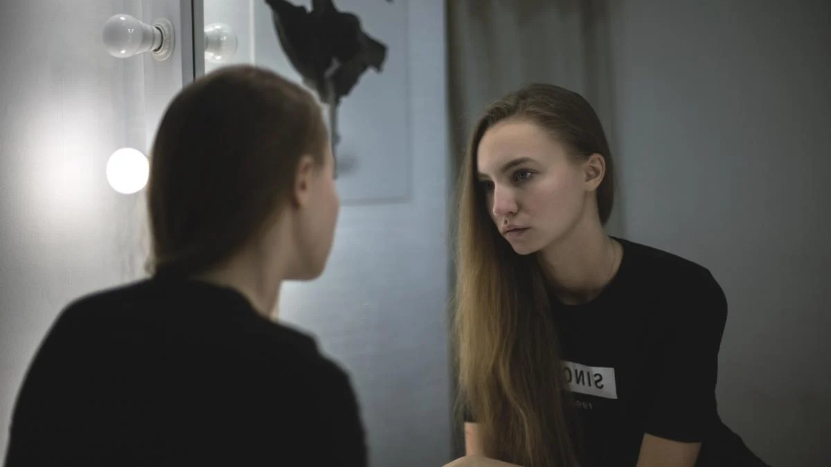 Young woman in a black shirt sits by a vanity mirror with round bulbs, gazing at her reflection.