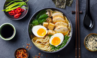 Ramen bowl topped with half-soft eggs, sliced chicken, greens, tofu cubes, and mushrooms on a dark table with sides of condiments are visible nearby.