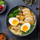 Ramen bowl topped with half-soft eggs, sliced chicken, greens, tofu cubes, and mushrooms on a dark table with sides of condiments are visible nearby.