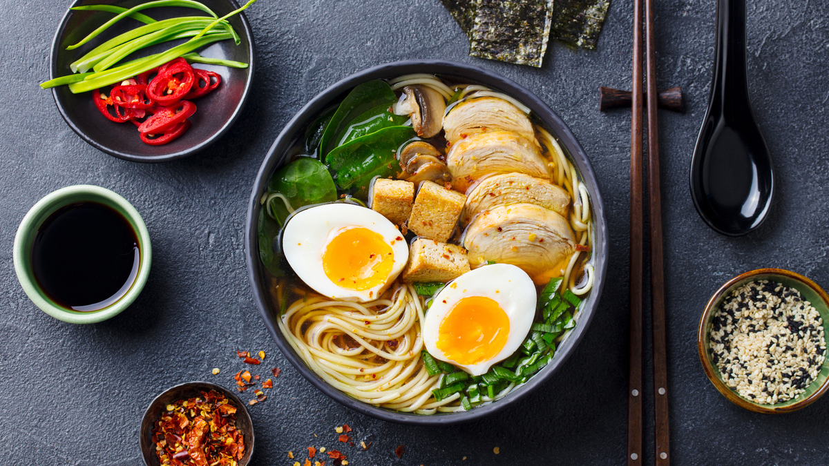 Ramen bowl topped with half-soft eggs, sliced chicken, greens, tofu cubes, and mushrooms on a dark table with sides of condiments are visible nearby.