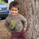 Smiling young boy in a beige dinosaur-print hoodie and maroon pants stands against a tree in a parking lot with cars behind him.