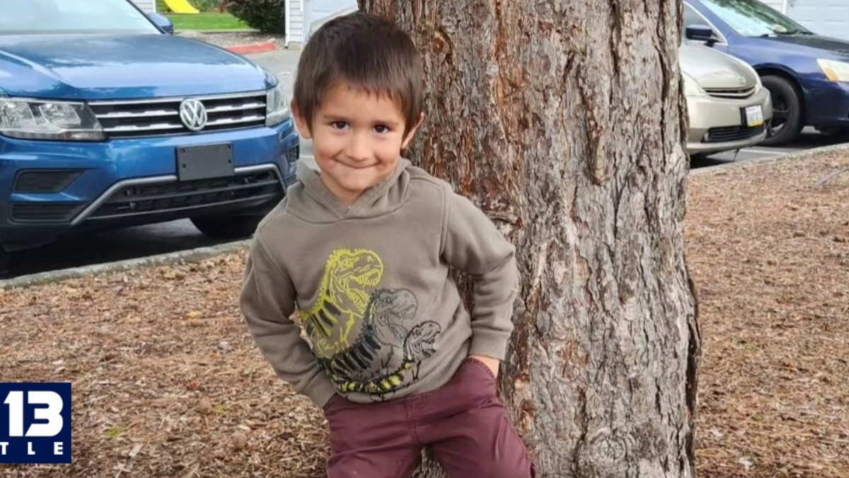 Smiling young boy in a beige dinosaur-print hoodie and maroon pants stands against a tree in a parking lot with cars behind him.
