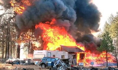 House and camper completely engulfed in bright flames with thick black smoke; a blue SUV and scattered debris sit in the foreground.