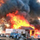 House and camper completely engulfed in bright flames with thick black smoke; a blue SUV and scattered debris sit in the foreground.