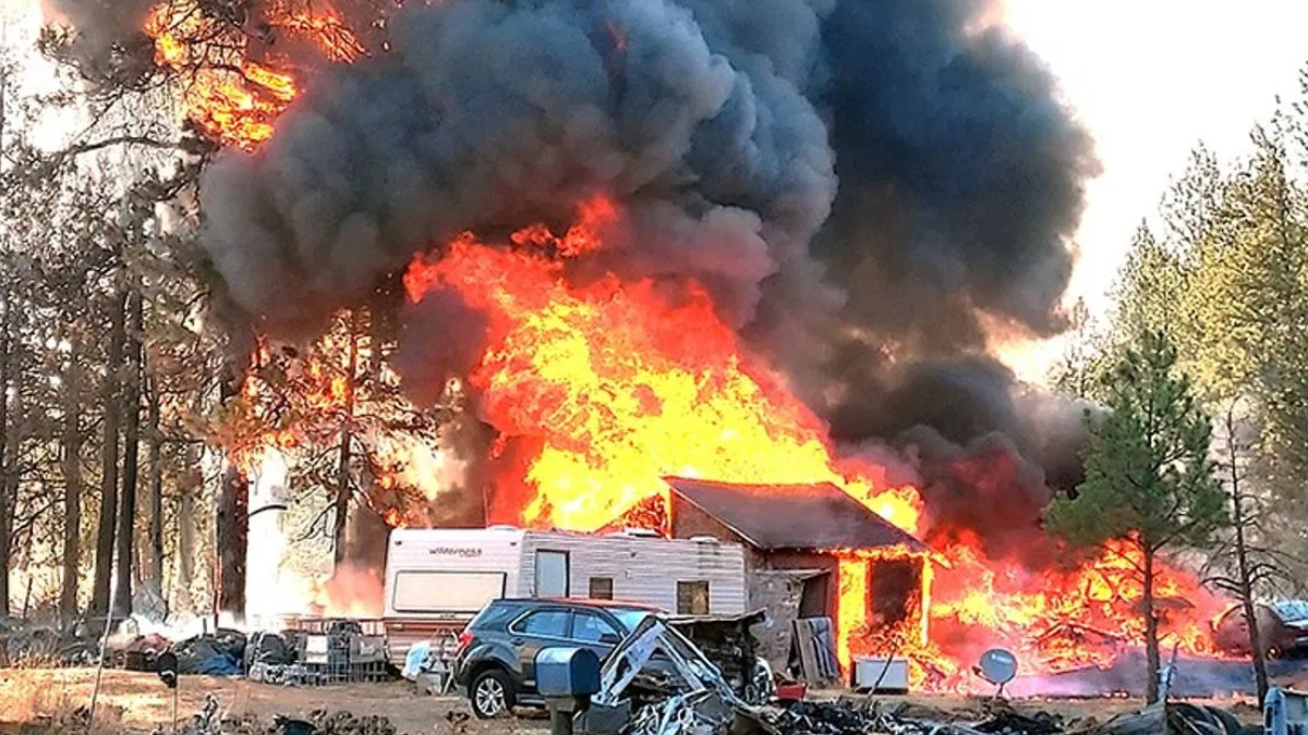 House and camper completely engulfed in bright flames with thick black smoke; a blue SUV and scattered debris sit in the foreground.