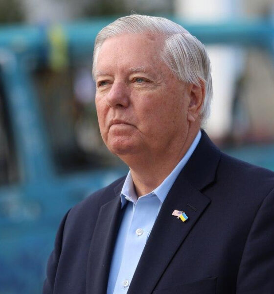 Serious man in a navy blazer and light blue shirt outdoors, wearing a small Ukrainian flag lapel pin, with a blue vehicle in the blurred background.