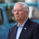 Serious man in a navy blazer and light blue shirt outdoors, wearing a small Ukrainian flag lapel pin, with a blue vehicle in the blurred background.