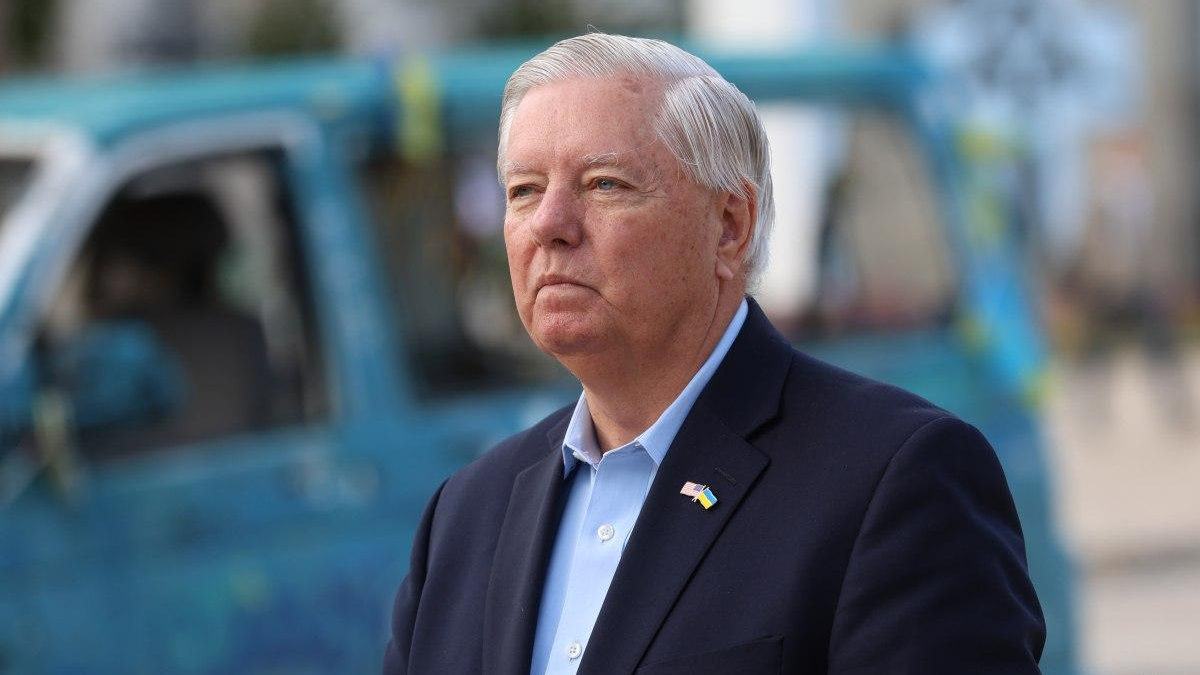 Serious man in a navy blazer and light blue shirt outdoors, wearing a small Ukrainian flag lapel pin, with a blue vehicle in the blurred background.