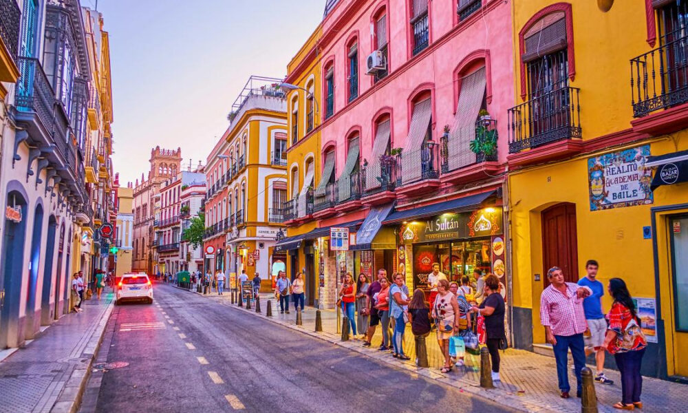 Colorful street in a European town with pink and yellow buildings and people queuing outside shops.