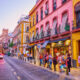 Colorful street in a European town with pink and yellow buildings and people queuing outside shops.