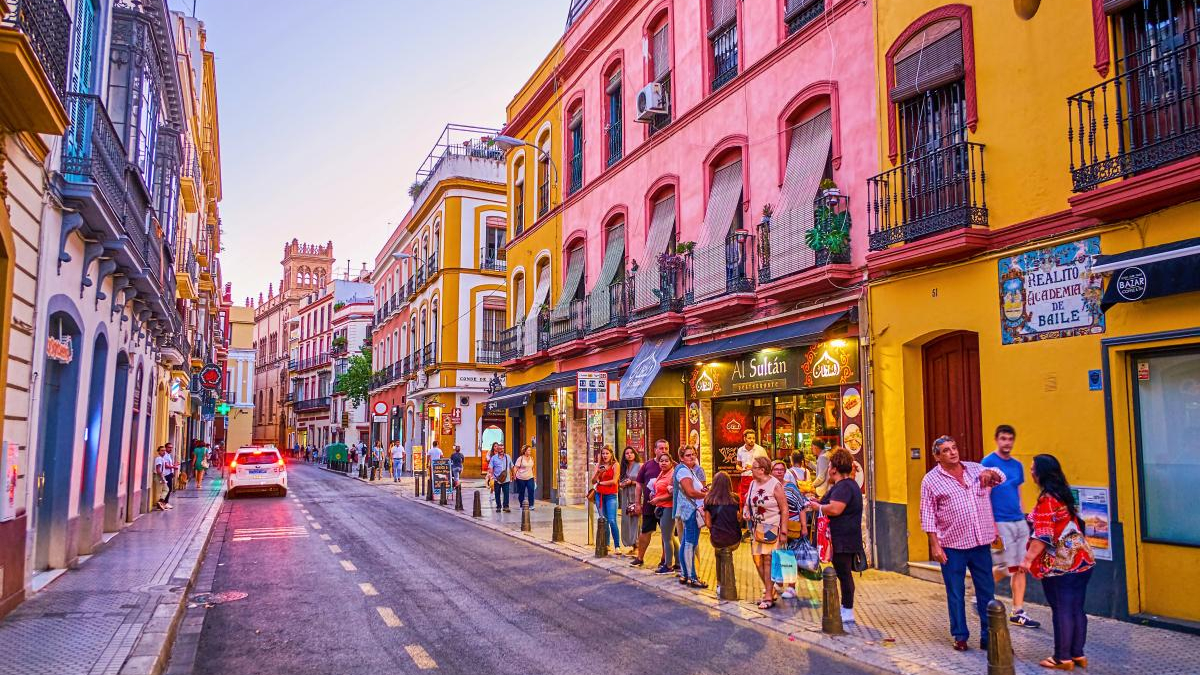 Colorful street in a European town with pink and yellow buildings and people queuing outside shops.