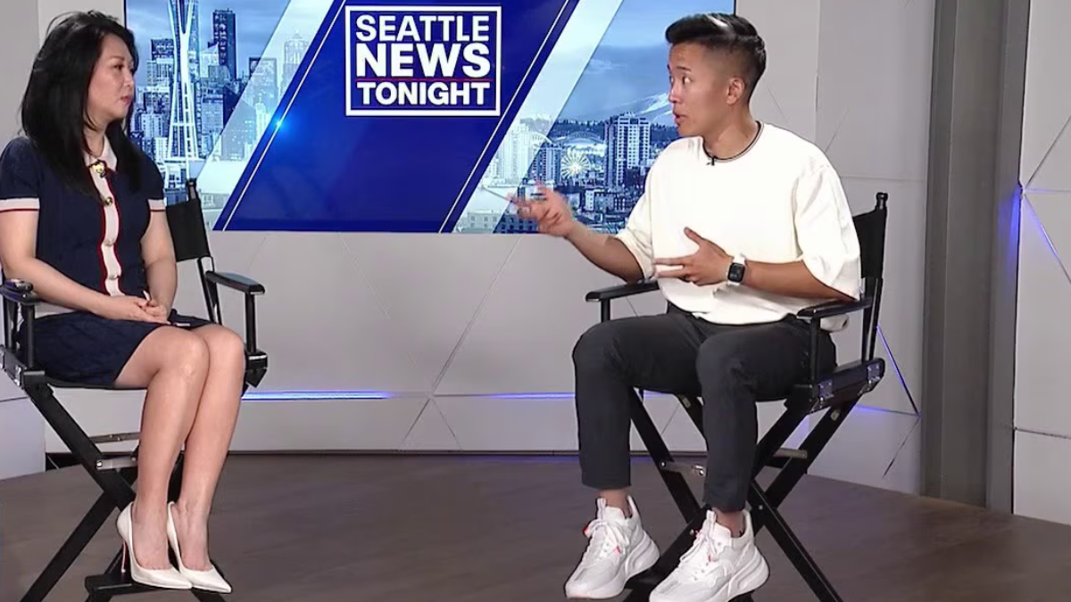 Two people sit in interview chairs facing each other on a news set with a Seattle skyline logo behind them, mid-conversation.