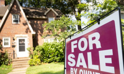 A large magenta 'FOR SALE' sign in the yard with a brick suburban house and greenery in the background.