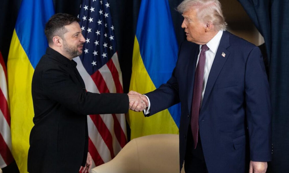 Two men in suits shake hands on a stage with American and Ukrainian flags behind them.