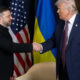Two men in suits shake hands on a stage with American and Ukrainian flags behind them.